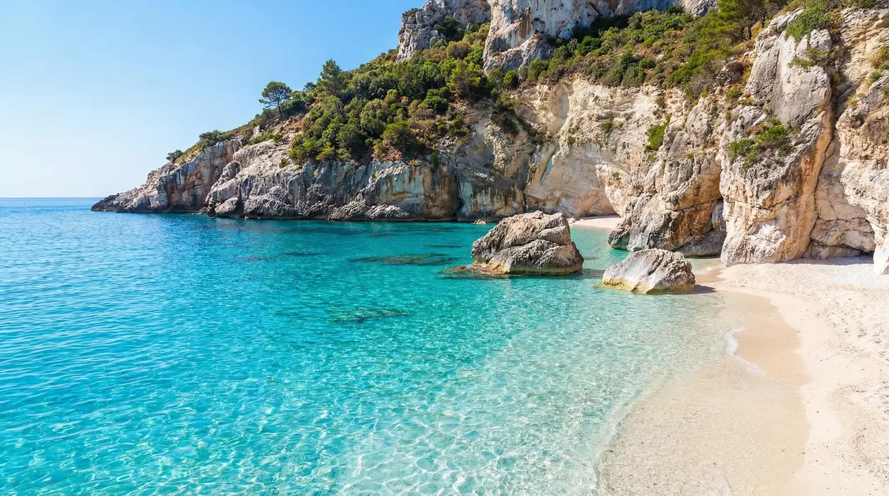 Cala con sabbia chiara, acqua turchese e scogliere rocciose ricoperte di vegetazione lungo la costa italiana