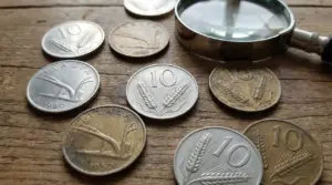 Vecchie monete italiane da 10 lire con spiga disposte su un tavolo di legno accanto a una lente d'ingrandimento.