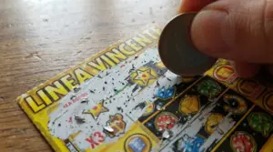 Mano che gratta un biglietto Gratta e Vinci con una moneta, mostrando i simboli del gioco sul tavolo