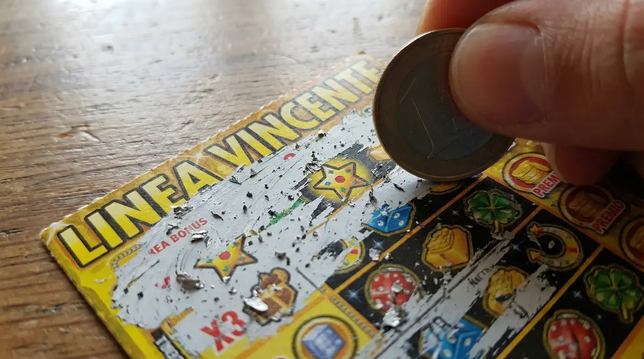 Mano che gratta un biglietto Gratta e Vinci con una moneta, mostrando i simboli del gioco sul tavolo