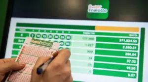 Mani che compilano una schedina del SuperEnalotto davanti a uno schermo con numeri e quote della vincita