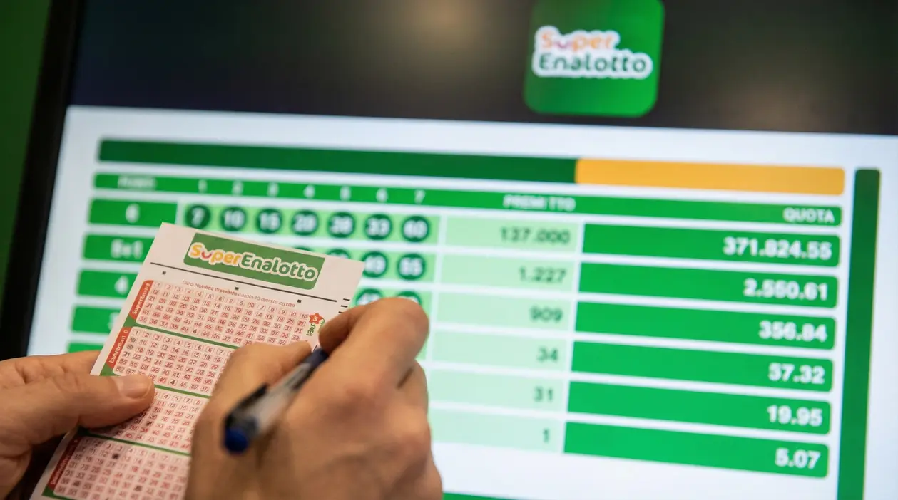 Mani che compilano una schedina del SuperEnalotto davanti a uno schermo con numeri e quote della vincita